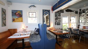 Een restaurant met houten tafels en blauwe zittingen met kussens, ingelijste foto’s aan de muren en een nautisch thema met vissen decoraties.