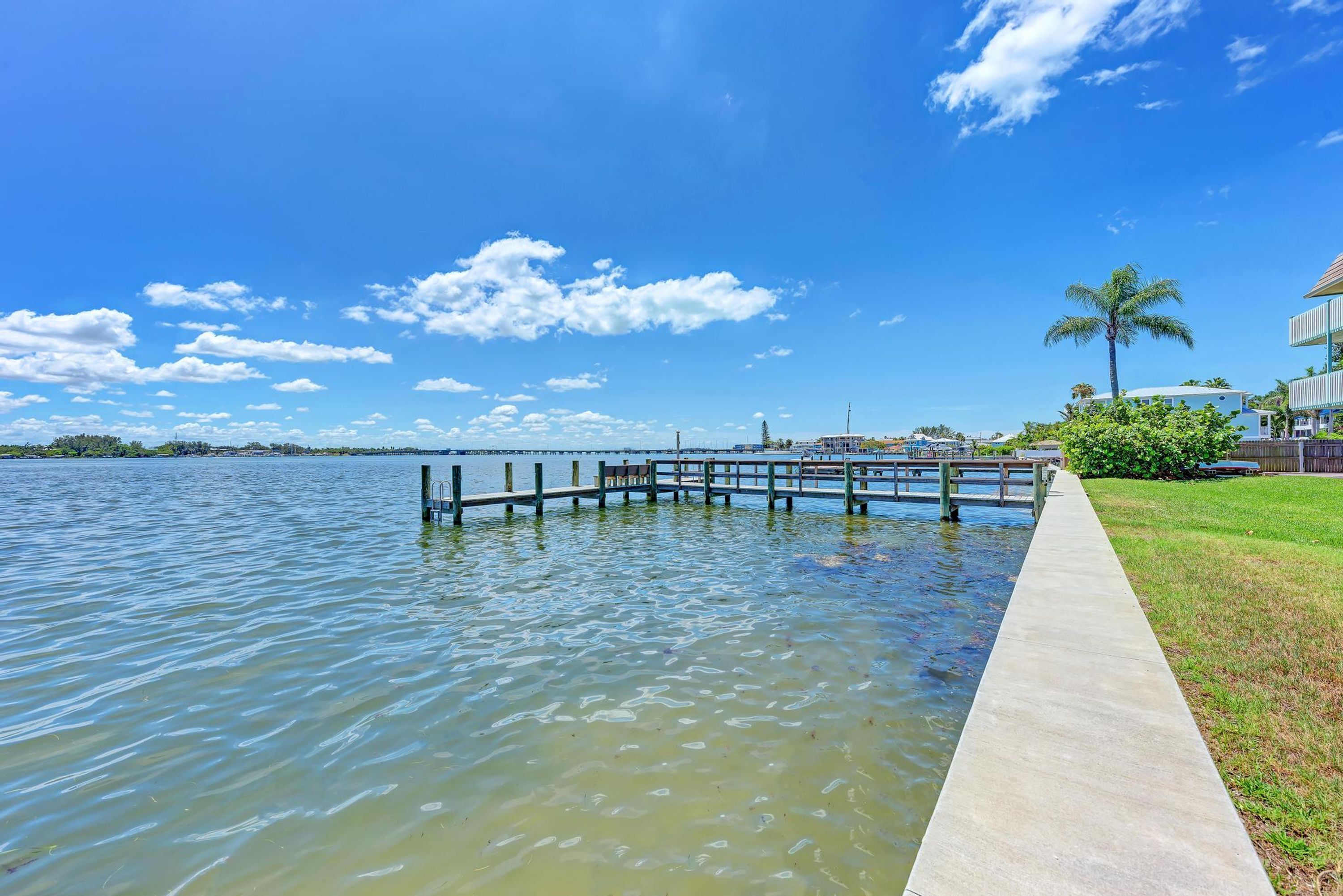 A waterfront walkway with a concrete path, a wooden dock extending into a calm body of water, and a clear blue sky with scattered clouds.