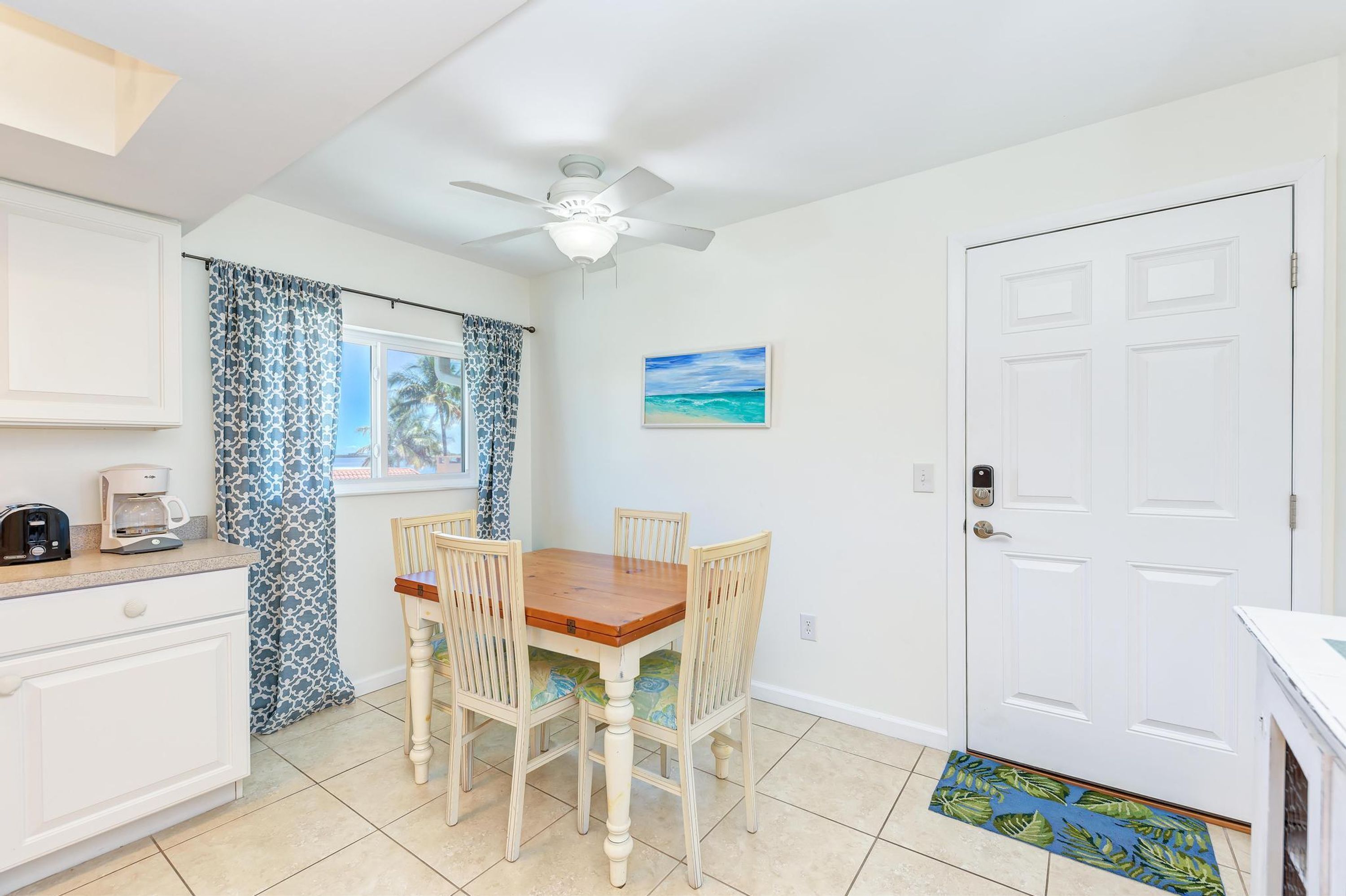 A dining area with a wooden table and four chairs, a ceiling fan, a window with curtains, a painting, and a white door.