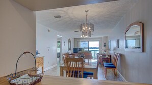 A spacious living area with a dining table, chairs, and a chandelier. There is a view of a balcony with a railing and a window. - Anna Maria Island Beach Palms 2A (Bradenton Beach)