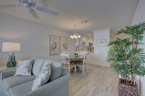 A modern living room with a grey sofa, a dining area with white chairs, and a kitchen in the background. - Anna Maria Island Beach Palms 2B (Bradenton Beach)
