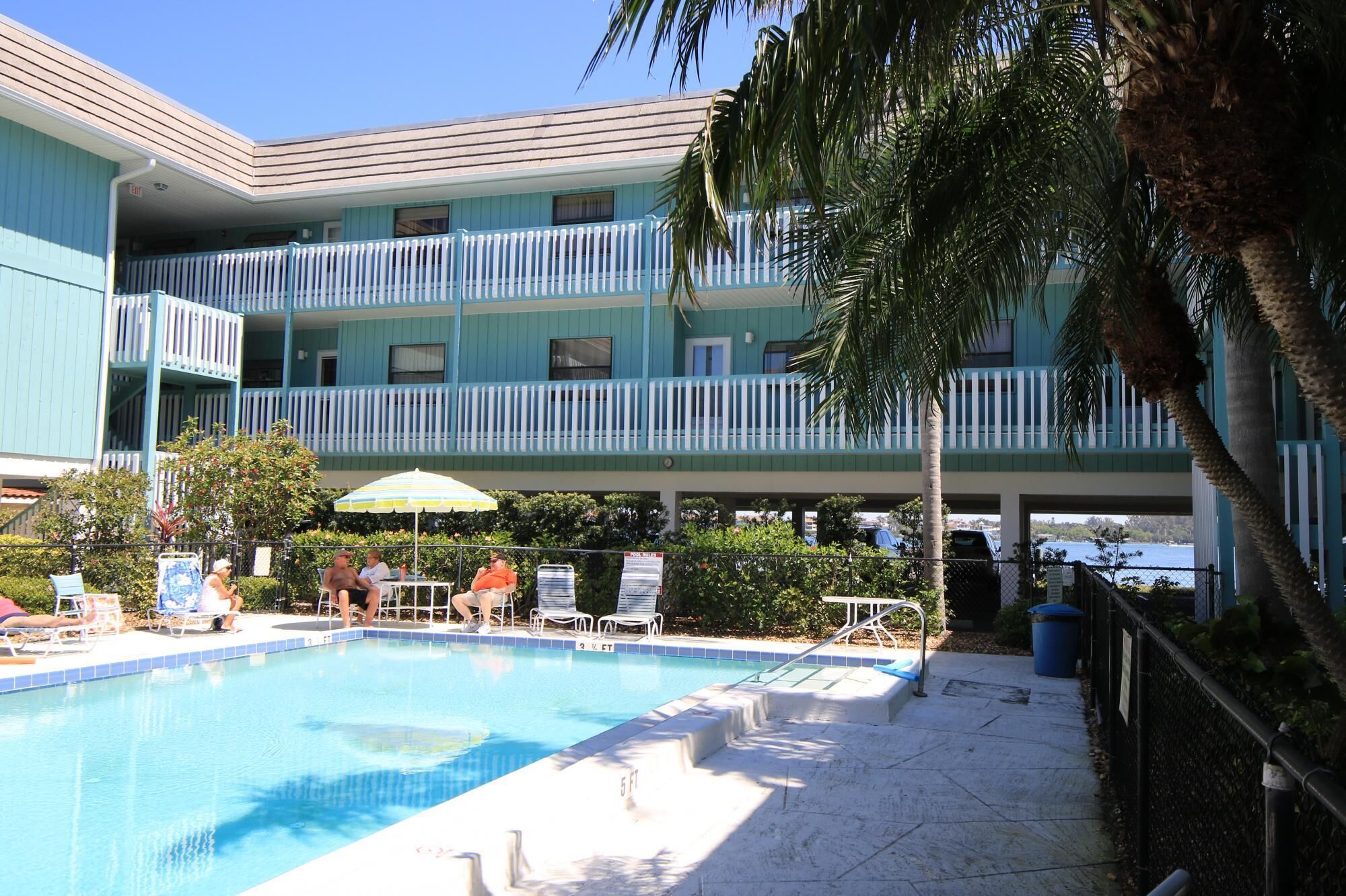 A pool area with lounge chairs and umbrellas, a multi-story building with balconies, and a clear view of the water.
