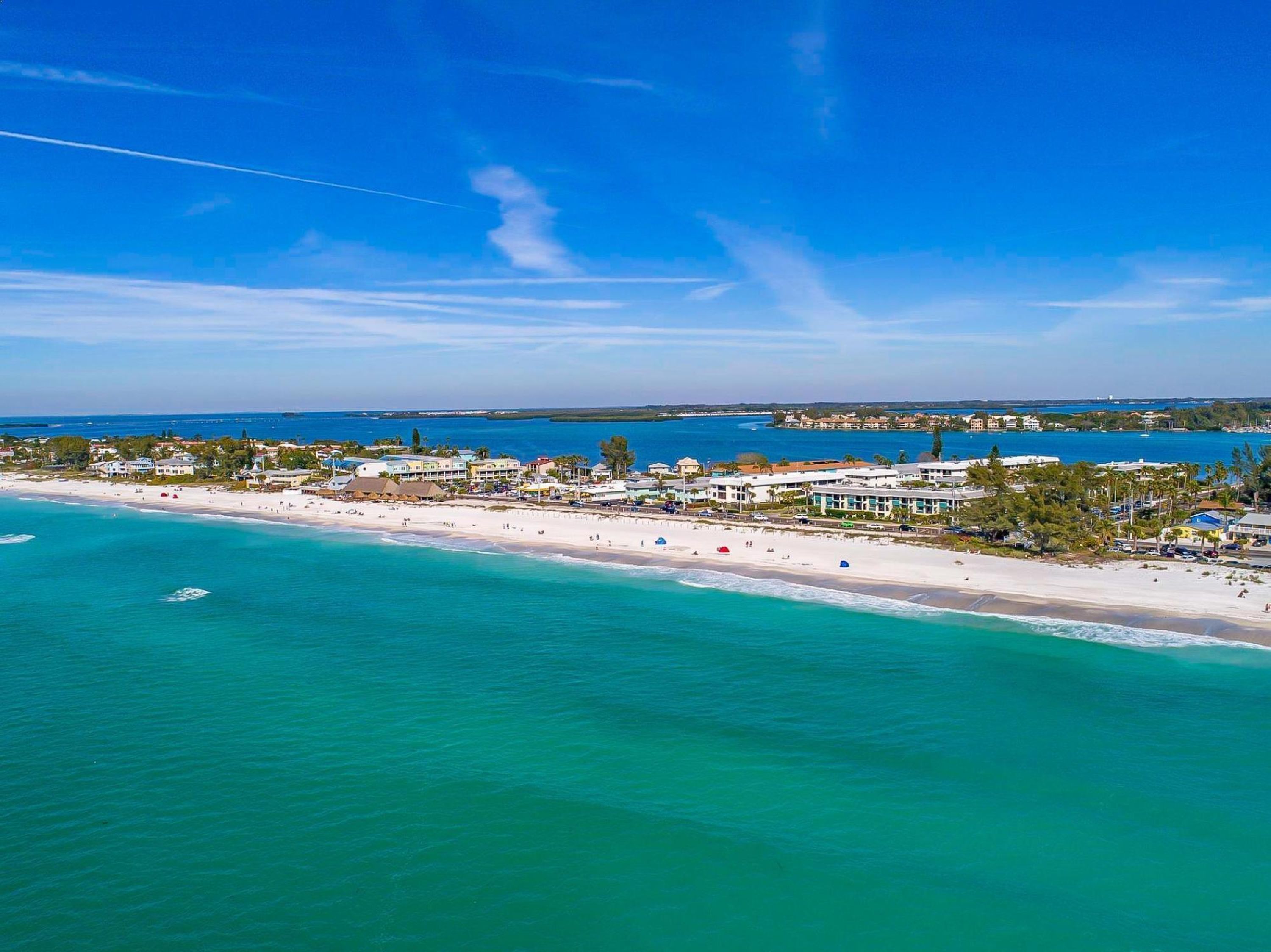 An aerial view of a coastal area with a beach, buildings, and clear blue waters.