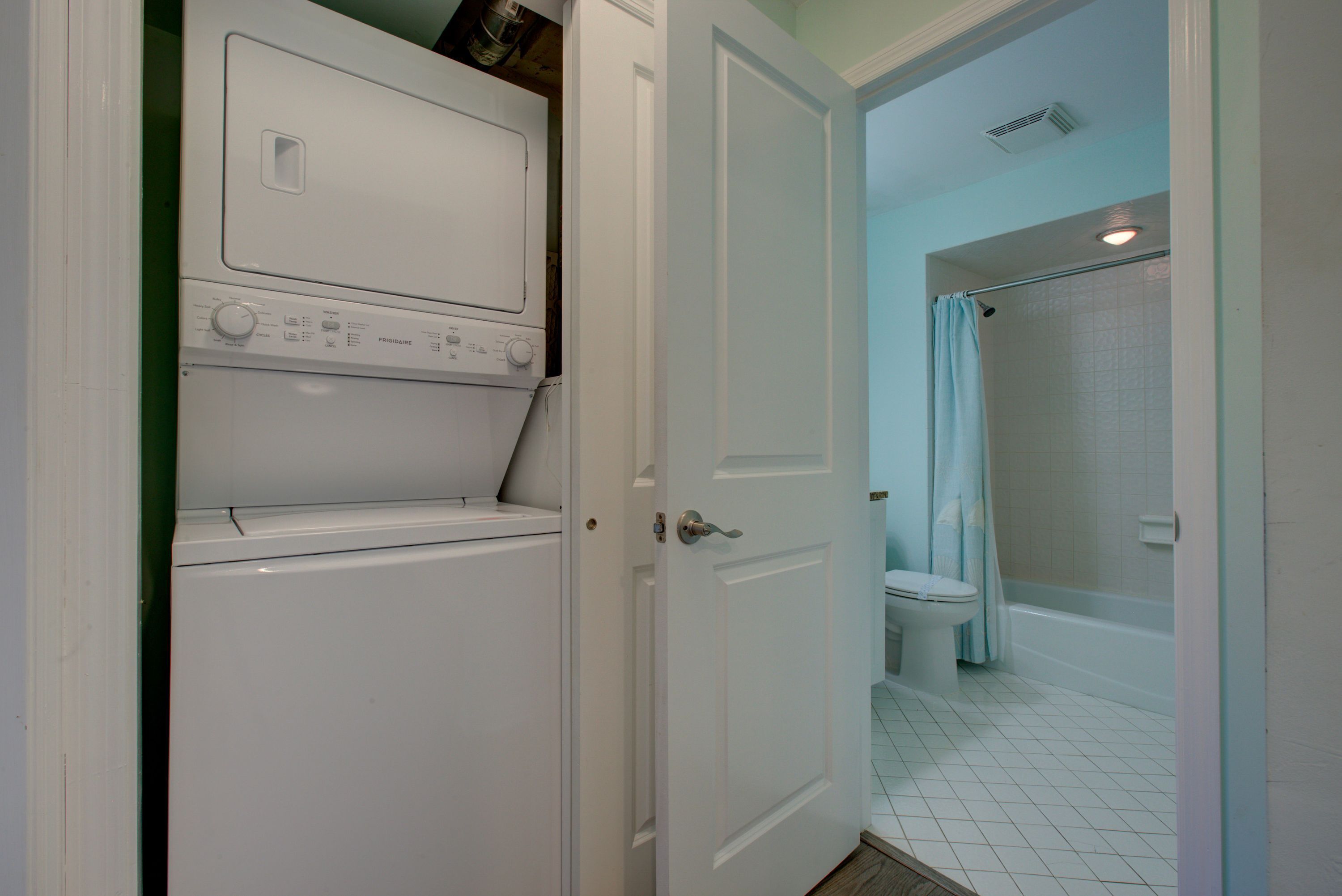 A laundry room with a front-loading washer and dryer, a bathroom with a toilet and shower, and a door leading to another room.