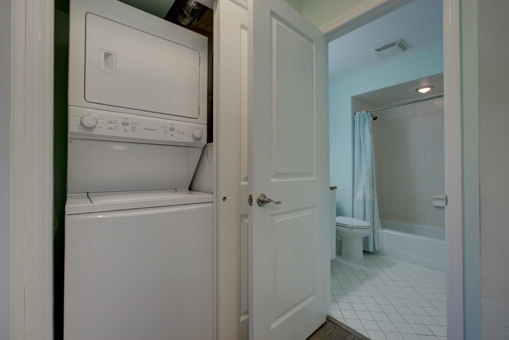 A laundry room with a front-loading washer and dryer, a bathroom with a toilet and shower, and a door leading to another room.