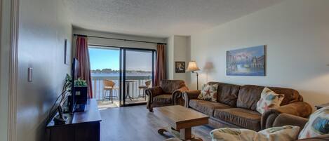 Een woonkamer met een bank, een salontafel en een televisie op een standaard. Er is een balkon met uitzicht op het water.
