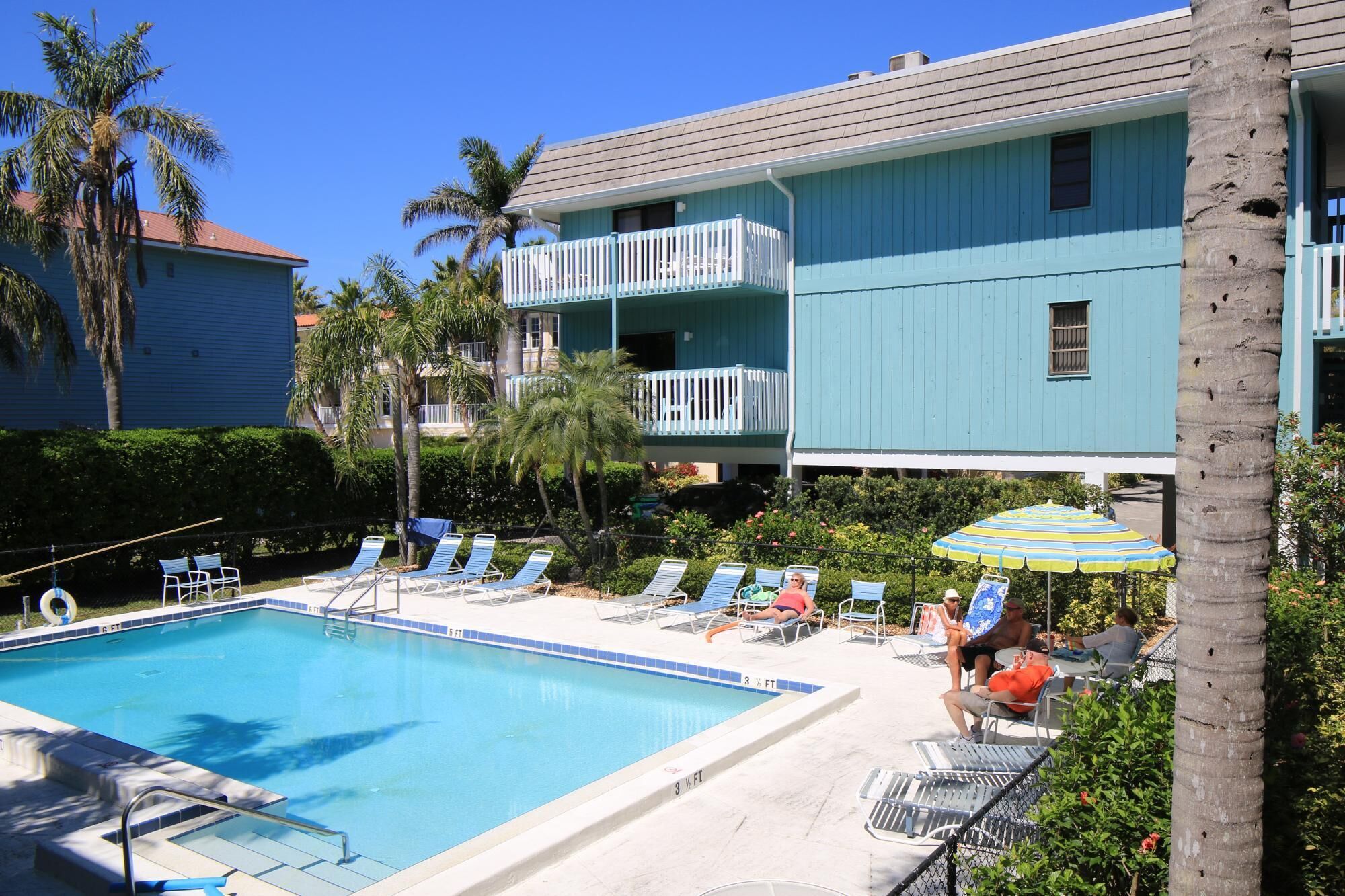 A swimming pool with lounge chairs and umbrellas, surrounded by palm trees and a two-story building with balconies.