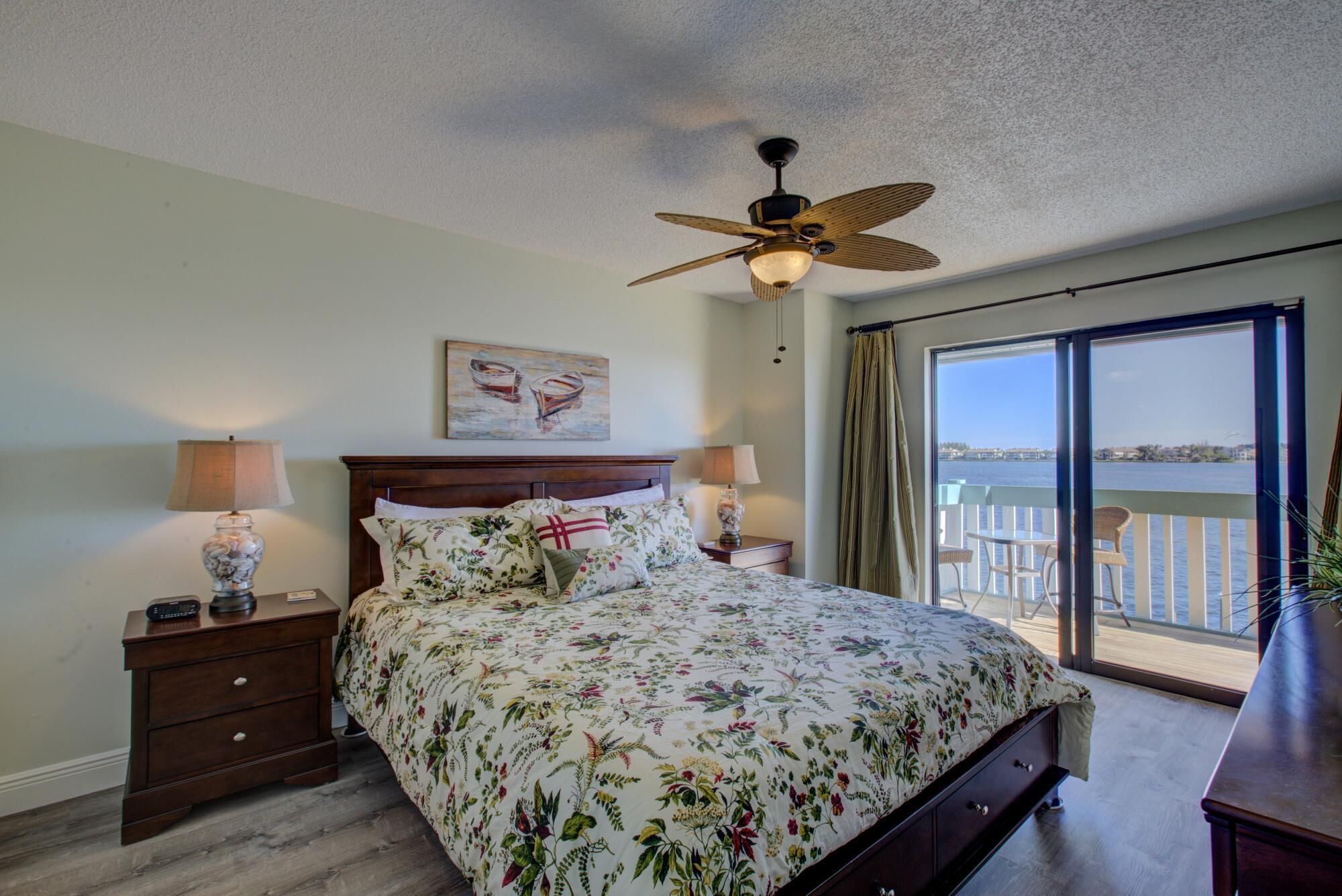 A bedroom with a bed, two bedside tables, a ceiling fan, a painting, and a view of the water through a large window.