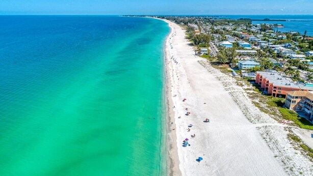 Luftaufnahme eines Strandes mit einer Reihe von Hotels direkt am Strand, klarem türkisblauem Wasser und einem langen Sandstrand.