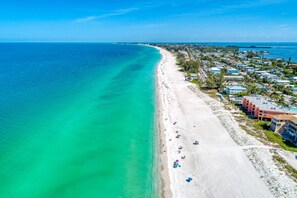 An aerial view of a beach with a row of beachfront hotels, clear turquoise waters, and a long sandy shore. - Anna Maria Island Beach Palms 6B (Bradenton Beach)