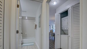 Un couloir mène à une salle de bain avec douche, à un espace buanderie équipé d’un lave-linge et d’un sèche-linge, et à un coin repas visible à travers une porte ouverte.
