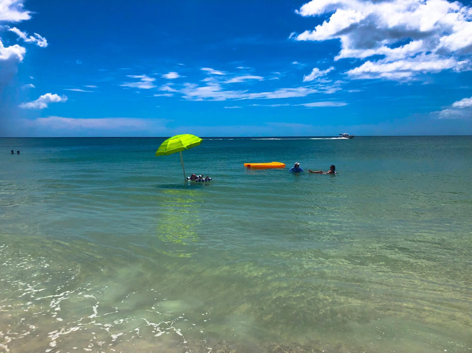 A clear blue sea with a person floating on a yellow raft under a green umbrella.