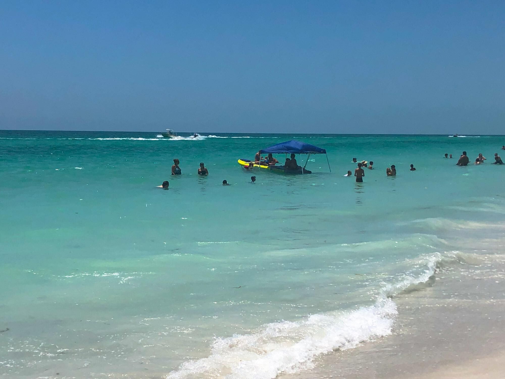 A beach with clear turquoise water, people swimming and a boat with a blue canopy.