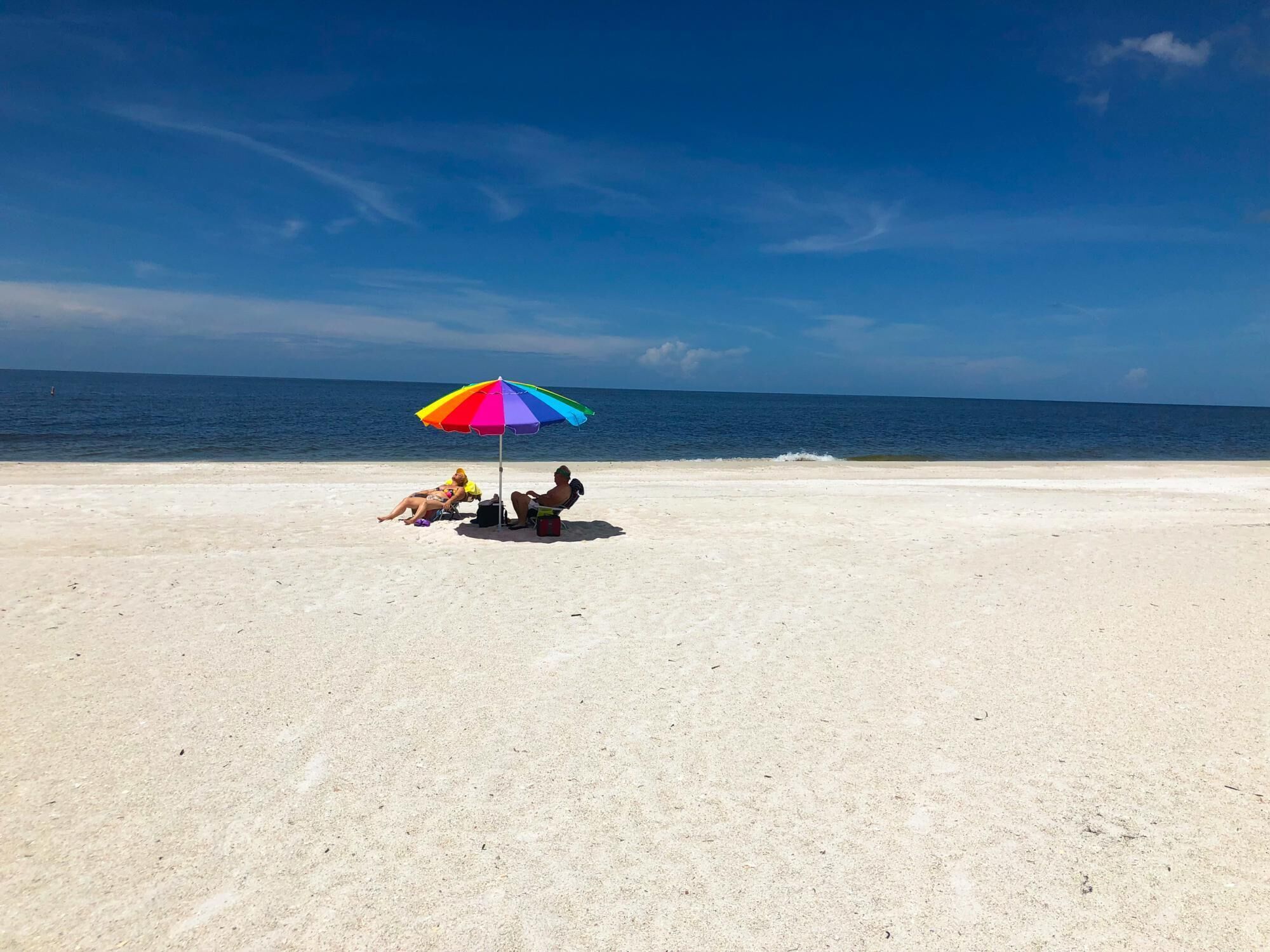 A beach scene with a colorful umbrella, two people relaxing under it, and the ocean in the background.