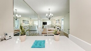 A modern living room with a large mirror, a dining area with a wooden table and white chairs, and a kitchen in the background.