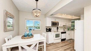 A modern kitchen with white cabinetry, a stainless steel microwave, and a white refrigerator. There is a dining table with chairs and a chandelier above.