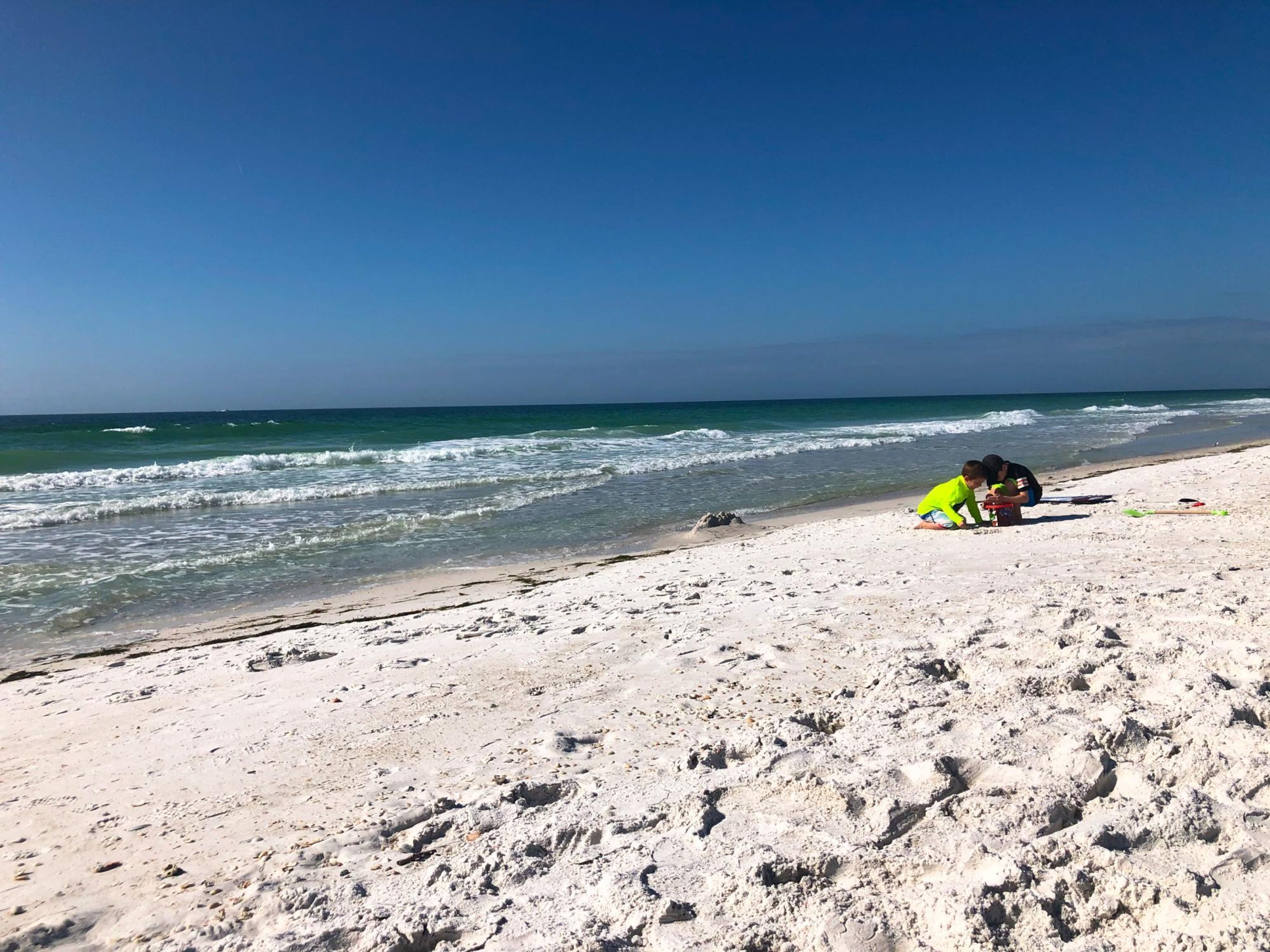 Een zandstrand met golven, twee personen zitten in het zand, en een helderblauwe lucht.