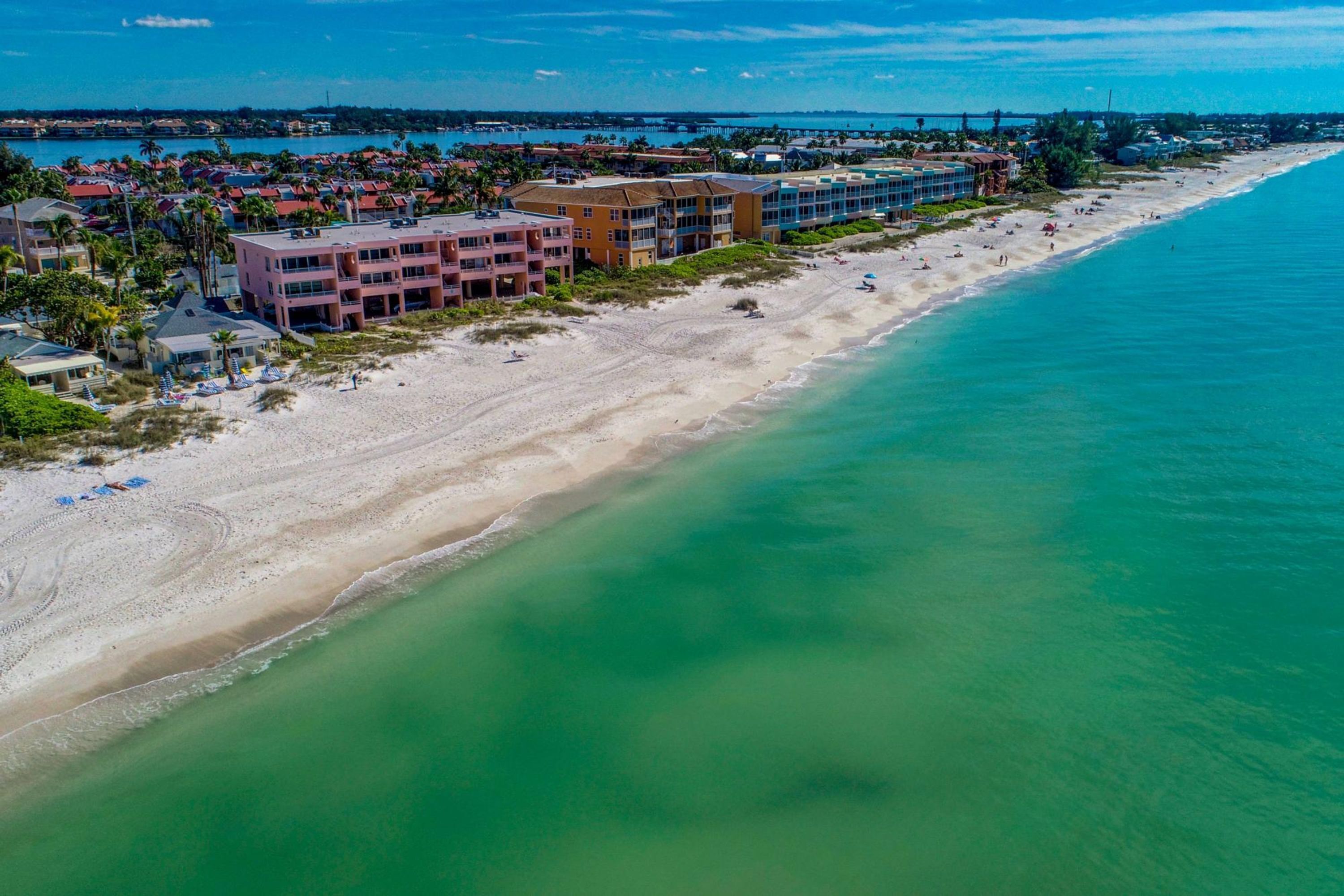 An aerial view of a coastal hotel with multiple buildings, a sandy beach, and a clear blue sea.