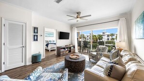 A spacious living room with a large window, a ceiling fan, a sofa set, and a television. - Modern 2br/2ba, Pet friendly, steps to private beach access and restaurants! (Miramar Beach)