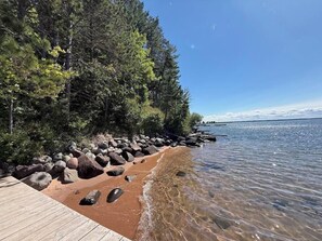 A wooden dock leading to a rocky shoreline with clear water and a forested area.