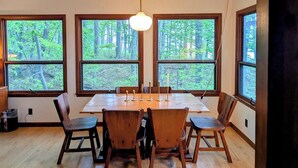 Un espace repas comprenant une grande table et des chaises, de grandes fenêtres donnant sur la forêt et un luminaire suspendu.
