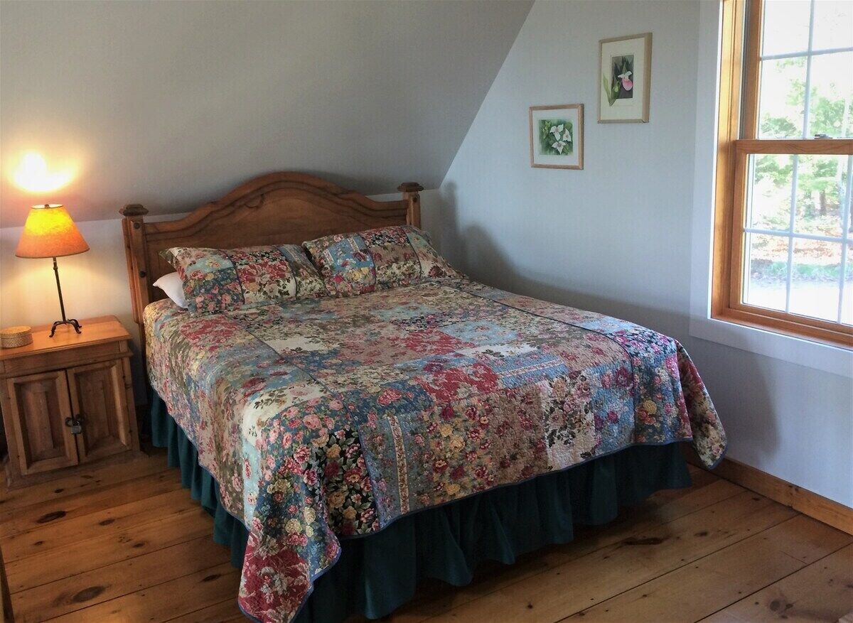 Un dormitorio con una cama de madera, una mesita de noche con lámpara, una ventana con vistas y una pared decorada con dos cuadros.