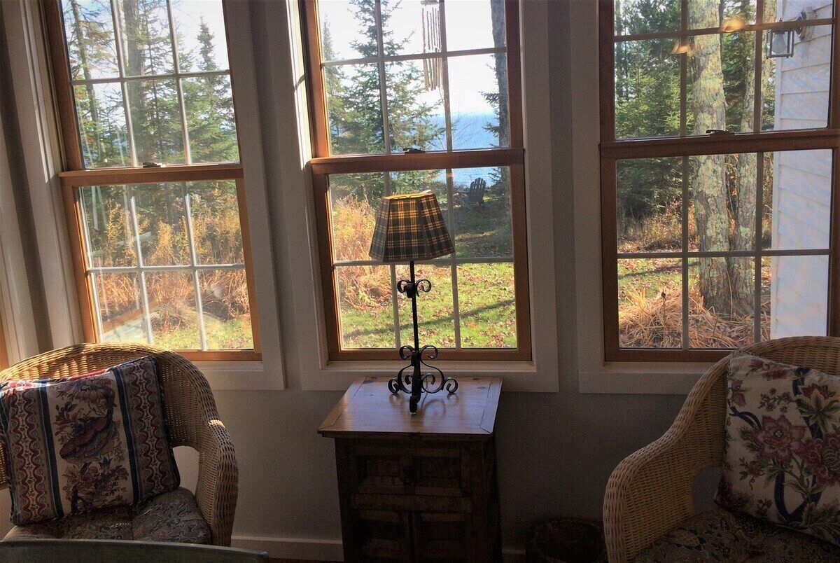 Una habitación con tres ventanas amplias, sillas de mimbre, una mesita con una lámpara y vistas a un bosque y al agua.