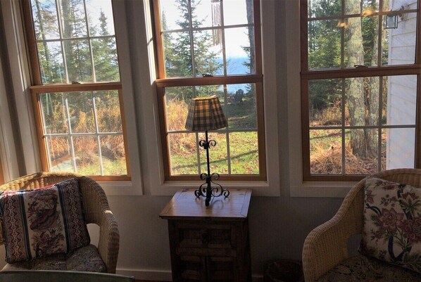 A room with three large windows, wicker chairs, a small table with a lamp, and a view of a forest and water.
