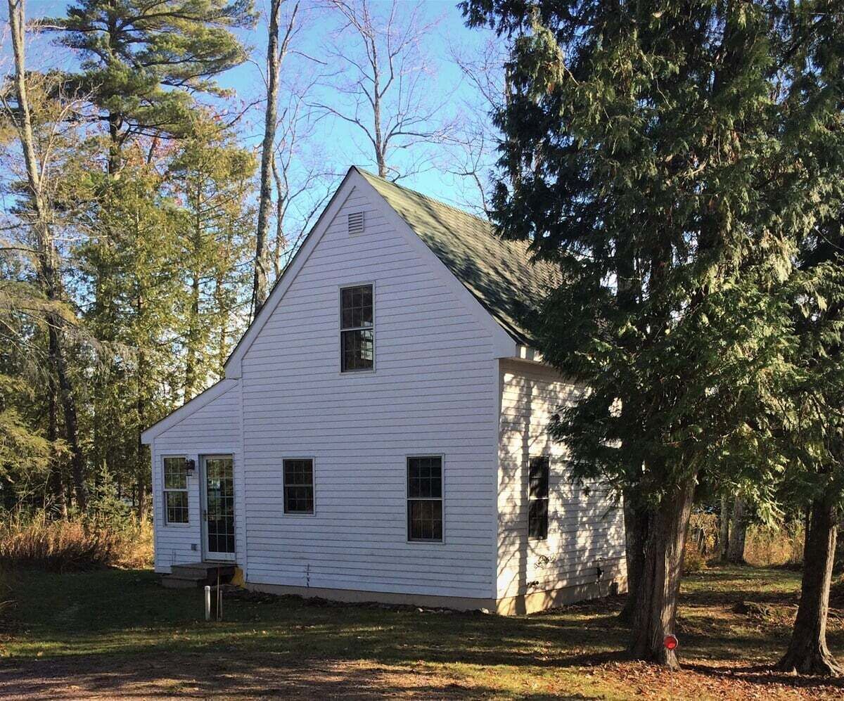 A white wooden house with a gabled roof, surrounded by trees and grass.