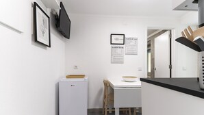 Una cocina compacta con un refrigerador blanco, una pequeña mesa de comedor con un plato, y una silla de madera. Hay cuadros en la pared y un menú colgado.