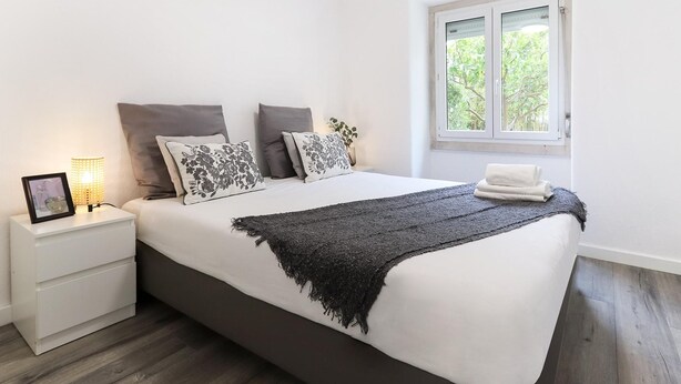 Un dormitorio con una cama grande, mesitas de noche blancas, una lámpara de noche y una ventana con vistas a la vegetación.