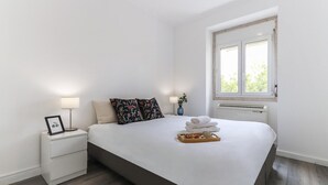 A neatly made bed with a tray of towels, a bedside table with a lamp, and a window with a view of greenery. - Belem Boutique VI by Homing (Lisbon)