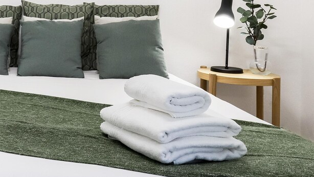 A neatly made bed with a green blanket, a stack of white towels, and a bedside lamp with a plant.