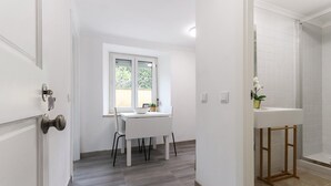 A hallway with a white door, a window with a view of greenery, a white table with chairs, and a bathroom with a sink and shower.