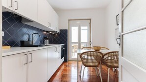 A narrow kitchen with white cabinets, a black backsplash, and a wooden floor. There is a dining area with wicker chairs and a table. - Belem Boutique IX by Homing (Lisbon)