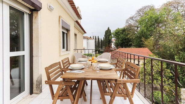 A wooden table set for four on a balcony with a railing, overlooking a garden with trees and a red-tiled roof building in the background. - Belem Boutique IX by Homing (Lisbon)