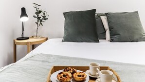 A neatly made bed with pillows, a bedside table with a lamp and a plant, and a tray with pastries and cups on a bedspread. - Belem Boutique II by Homing (Lisbon)