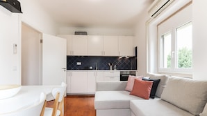 A compact kitchen with white cabinets, a black backsplash, and a built-in oven. There is a dining area with a white table and chairs, and a grey sofa with cushions. - Belem Boutique Viii by Homing (Lisbon)