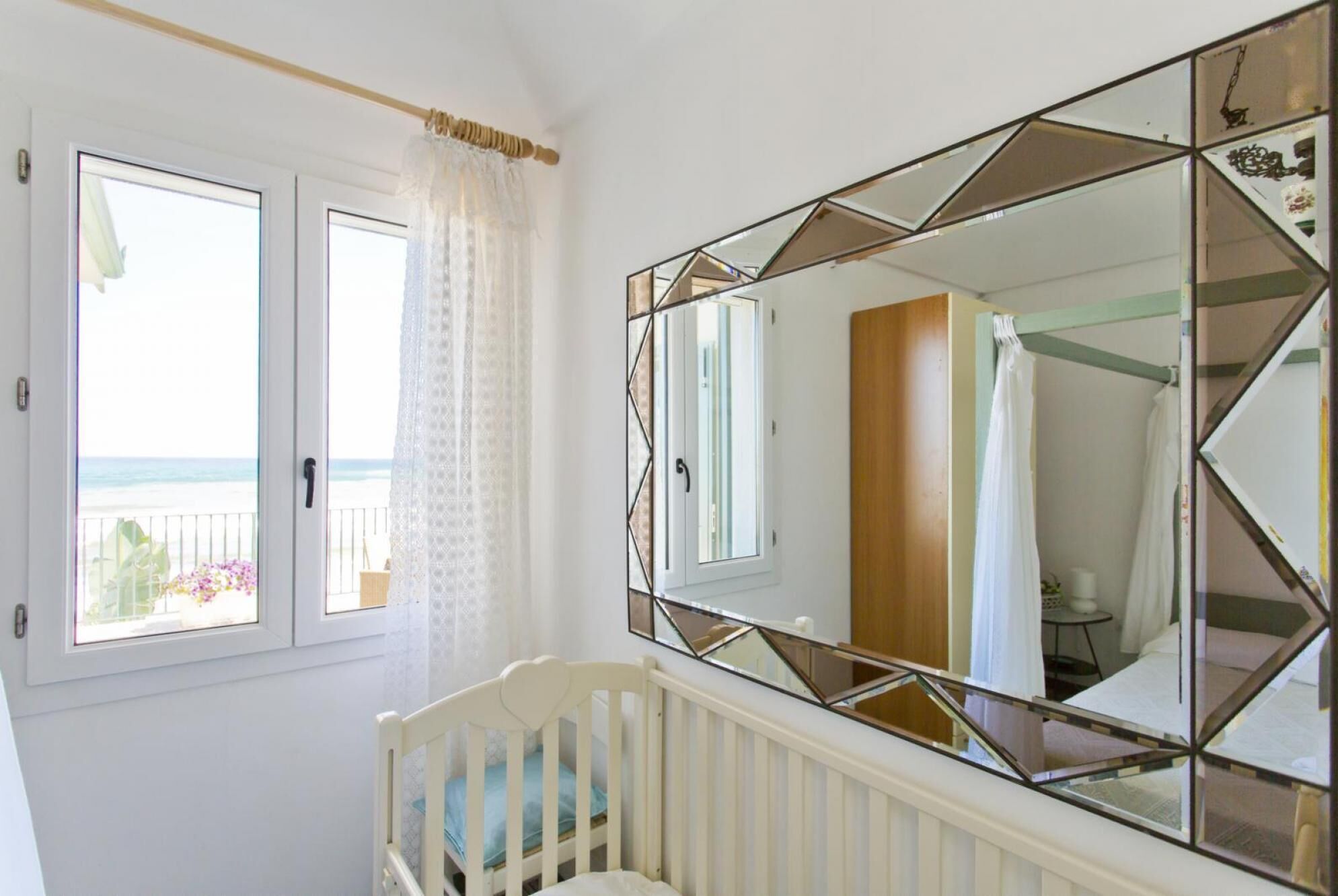 Quarto com berço, um espelho grande com padrões geométricos e janela com vista para a praia.