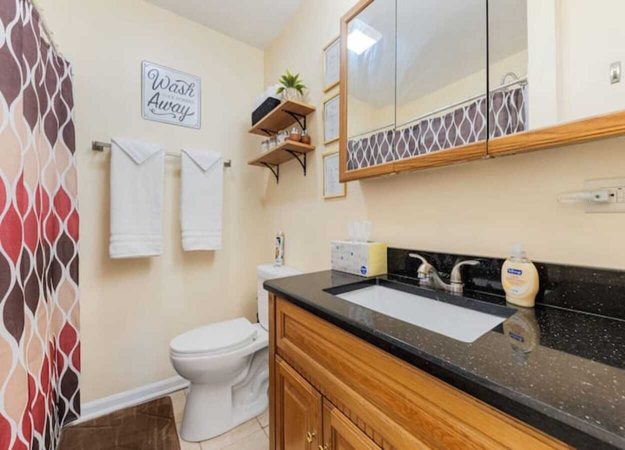 Ein Badezimmer mit Waschbecken, Spiegel und Toilette. Handtücher und ein Handtuchhalter, eine Duschvorhang und ein Schild mit der Aufschrift 'Wash Away'.