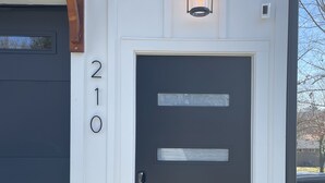 Una puerta negra moderna con tres ventanas rectangulares, una pérgola de madera encima y un farol iluminado.