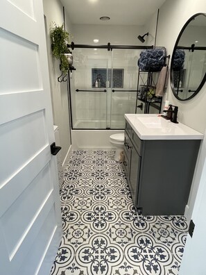A bathroom with a patterned floor, a shower, a toilet, and a sink with a mirror. - Farmhouse Loft Retreat (Knoxville)