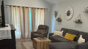 Una sala de estar con un sofá gris, un sillón marrón, una mesa de centro de madera y una consola blanca con un letrero de bienvenida.
