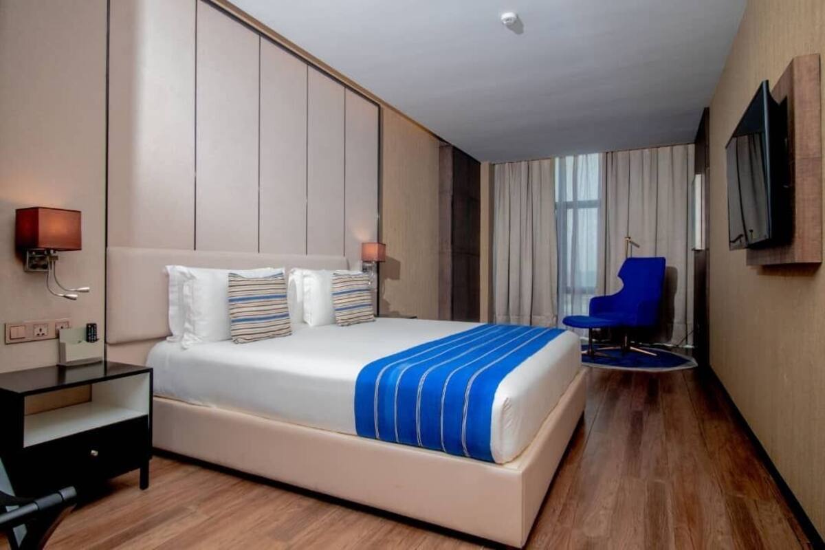 Quarto de hotel com cama grande, mesa de cabeceira, cadeira, televisão e janela com cortinas.