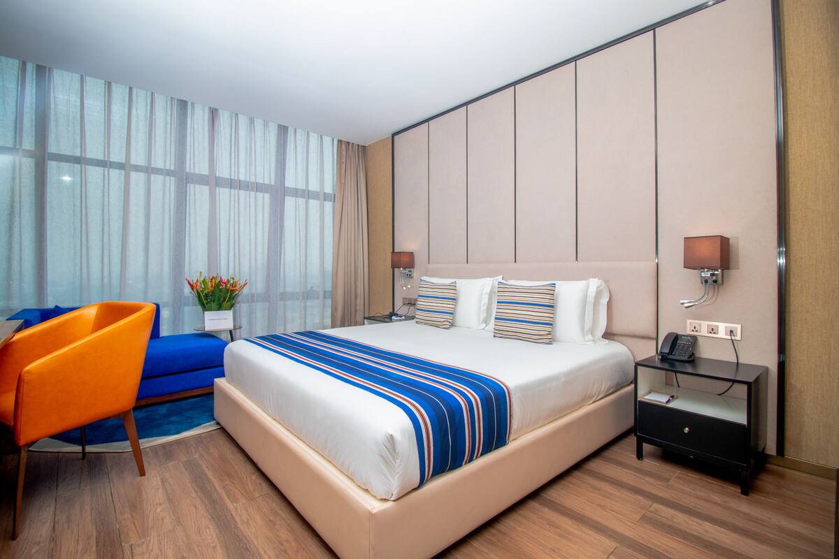 Quarto de hotel com uma cama grande, uma mesa de cabeceira, uma cadeira e uma janela com cortinas.