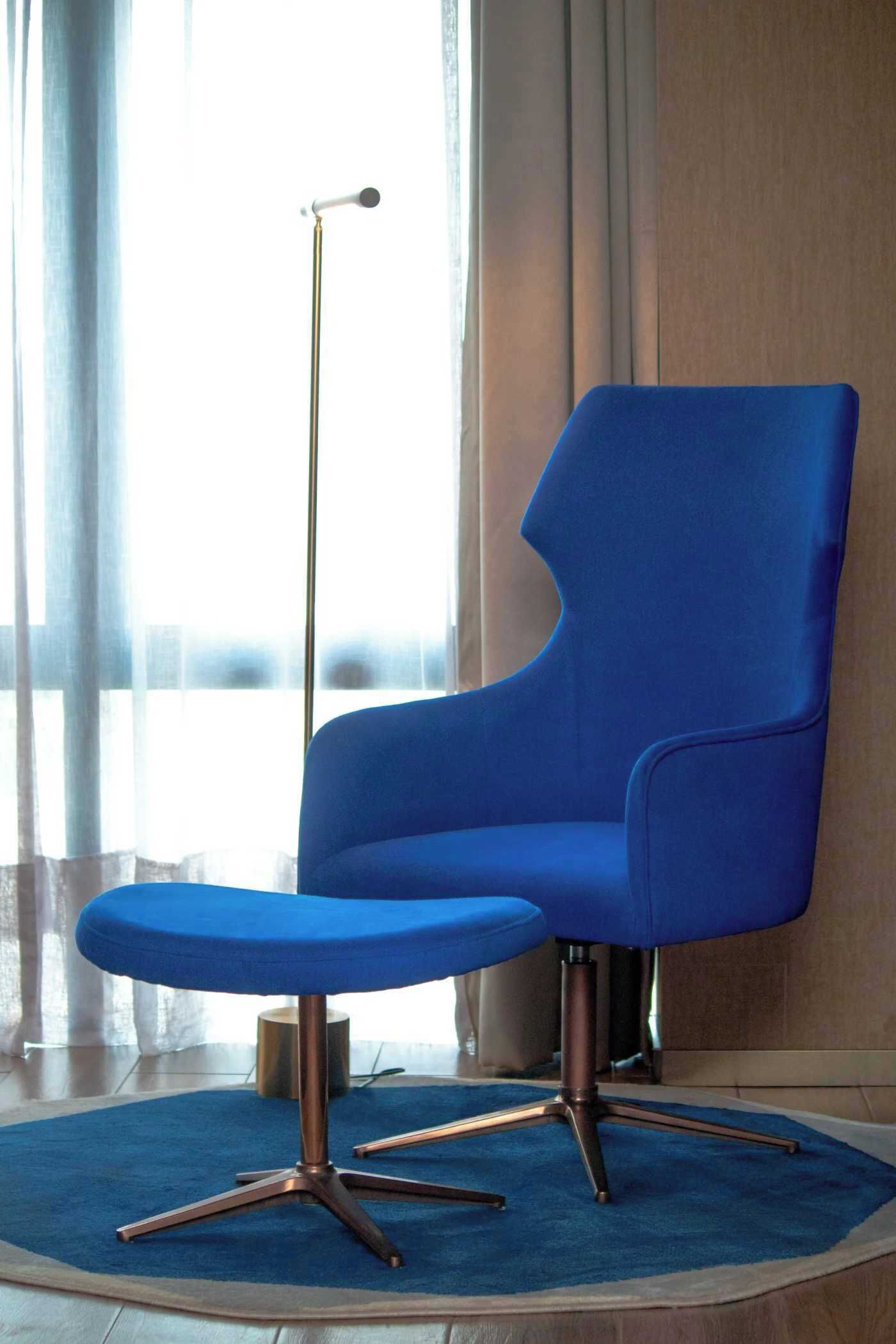 Un sillón azul con respaldo alto y un reposapiés a juego, sobre una alfombra azul en una habitación con suelo de madera y una ventana con cortinas de encaje.