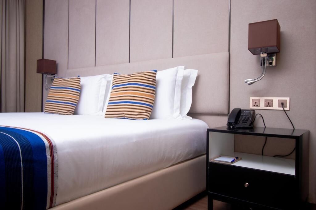 A hotel room with a bed, two pillows, a bedside table with a phone, and a wall-mounted light.