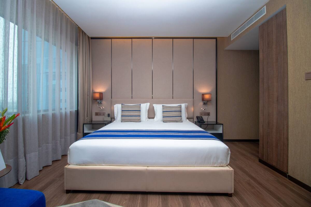 Quarto de hotel moderno com uma cama grande, duas mesas de cabeceira com luminárias, uma janela ampla com cortinas finas e piso de madeira.