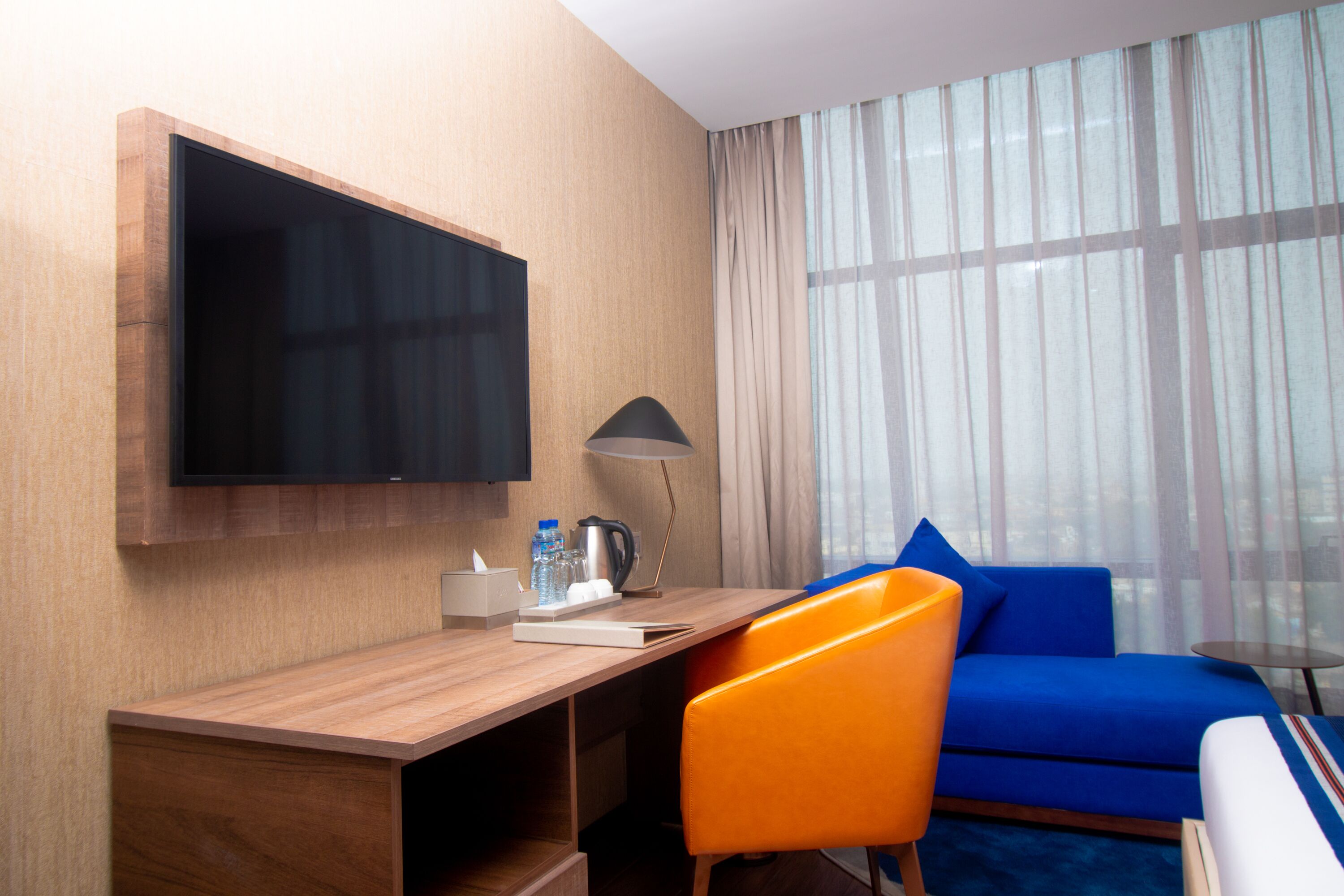 Habitación de hotel con un escritorio de madera, un televisor de pantalla plana, una lámpara, un sofá azul y vistas a través de cortinas de gasa.