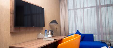 Habitación de hotel con un escritorio de madera, un televisor de pantalla plana, una lámpara, un sofá azul y vistas a través de cortinas de gasa.